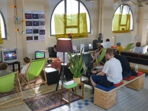 Menschen spielen Videospiele an alten Bildschirmen in einem Raum mit grünen Liegestühlen und Bogenfenstern.