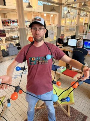 Mann im „Retro Computer Dresden“-T-Shirt umwickelt mit bunter Lichterkette aus großen Glühbirnen.