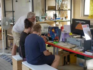 Drei Personen betrachten einen Vintage-Computer mit Grafik auf dem Monitor in einem Arbeitsraum.