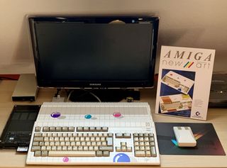 Ein Amiga 500 im „Ball-Design" steht einsatzbereit auf einem Schreibtisch vor einem modernen Samsung-Monitor. Rechts daneben steht ein aufgestellter Werbeflyer der „Amiga New Art Limited Edition". Zum Setup gehören außerdem eine klassische Commodore-Maus auf einem dunklen Mauspad sowie technische Erweiterungen auf der linken Seite.