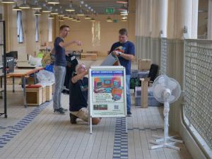 Drei Männer richten in einer Halle ein Schild für die Retro Computer Lounge auf.